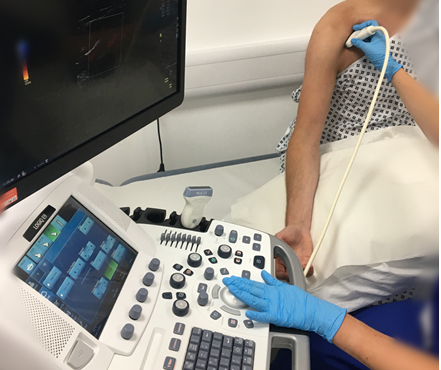 A patient sat on a hospital bed with a vascular scientist using an ultrasound machine to scan the patient’s shoulder region