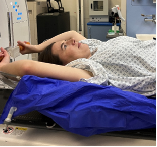 Person lying on an imaging table with arms raised above the head, wearing a hospital gown in a clinical scanning room.
