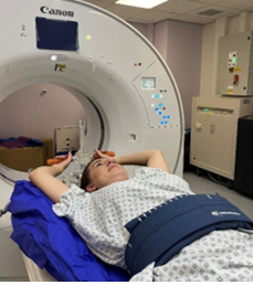 Person lying on an imaging table with arms raised above the head, wearing a hospital gown in a clinical scanning room.