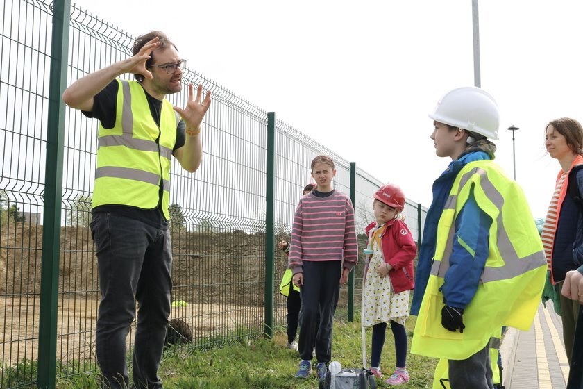 A group of children, with some wearing builders hard hats, listening to a man who is animatedly explaining about the archeological dig going on behind him, on the Cambridge Children's Hospital site during pre construction works