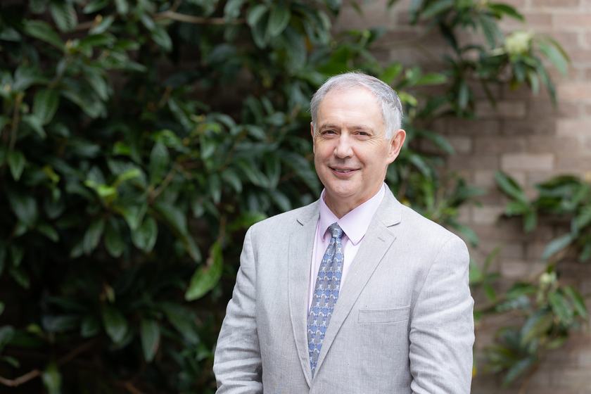 A man with grey hair, a grey suit, pink shirt and pale blue tie, standing outside