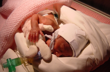 Baby with eyes closed, holding a soft toy, laying in an intensive care hospital cot bed with a pink waffle cover. The baby has a tube going into its mouth.