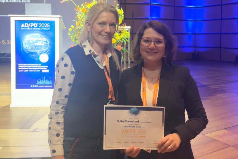 Caroline Williams-Gray and Julia Greenland next to each other in front of the stage at the ADPD 2025 conference. Julia is holding her junior faculty award certificate