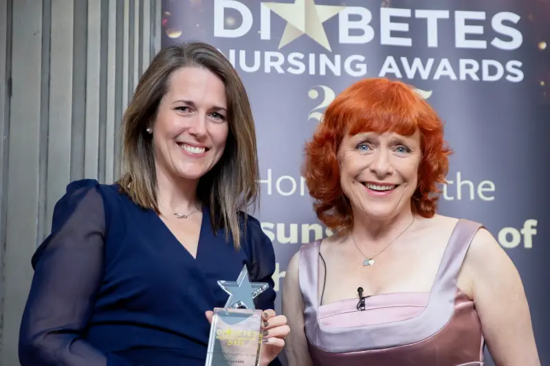 Andrea Lake and Dr Sarah Jarvis standing together in front of a banner for the Diabetes Nursing Awards. Andrea is holding her star-topped transparent trophy.