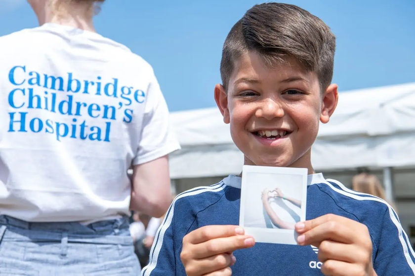 A young boy holding a polaroid photo of two hands creating a heart shape, during a workshop at the CUH Arts festival. The back of a person can be seen behind him with 'Cambridge Children's Hospital' printed on their T-shirt