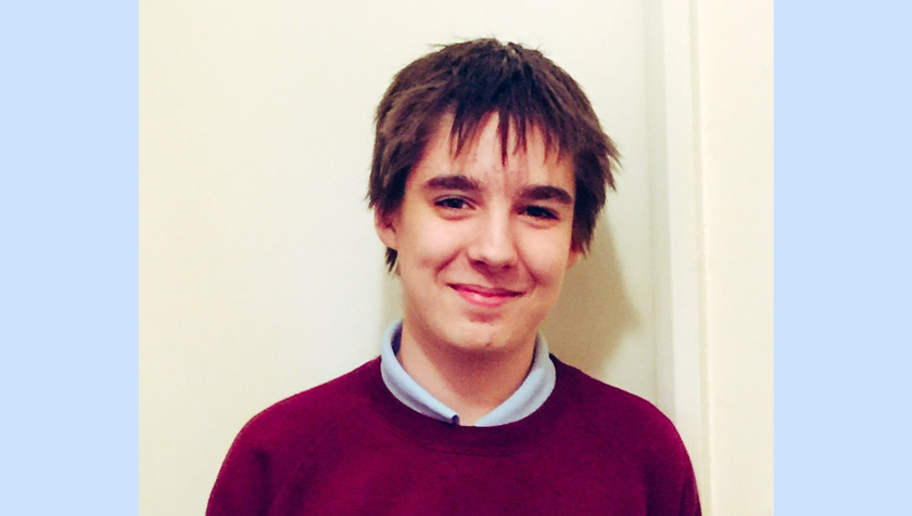 A boy with dark brown messy hair in a maroon school jumper. He is smiling