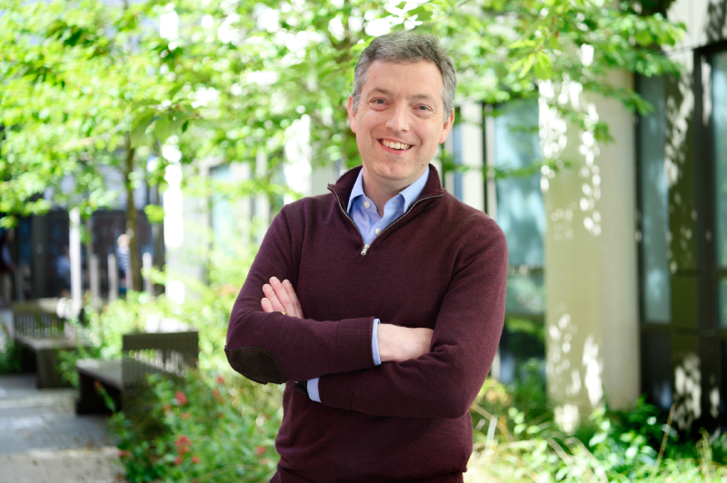 Ben standing in a relaxed pose with his arms crossed outside in front of a building and trees.