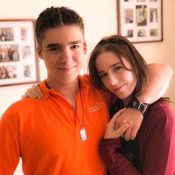 A woman with long dark hair standing with her son who has short dark hair and a red t shirt. He has his arm around her