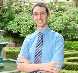 Dr Brent O'Carrigan - He has brunette hair, wears a blue shirt and striped tie and is standing outdoors with a fountain behind him.
