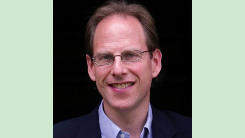 A man with brown slightly receding hair and rectangular rimmed glasses. He is wearing a pale blue shirt and suit jacket