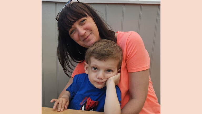 A little boy with brown hair sitting at a table leaning on his elbow. He's on his mother's lap. She has long very dark hair and is wrapping her arms around him.