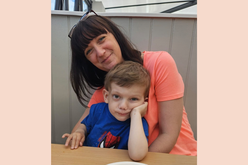 A little boy with brown hair sitting at a table leaning on his elbow. He's on his mother's lap. She has long very dark hair and is wrapping her arms around him.