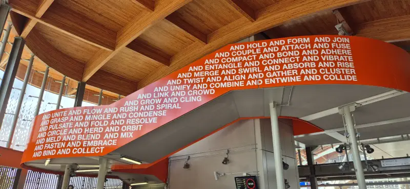 A raised section of stairway under a wooden roof. The side of the stairs is painted orange and covered in many written words, making up the artwork.
