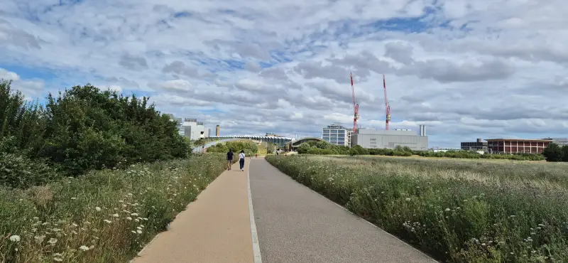 A straight pathway leading to the station with the Biomedical Campus in the background. The station is almost hidden by surrounding plants and trees.