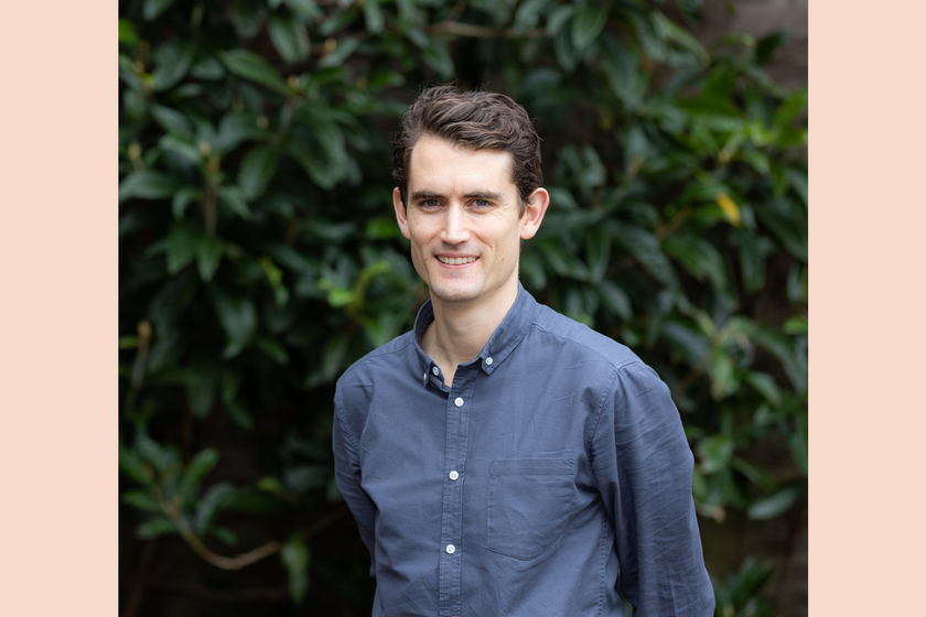 A man with black hair, short and combed to the side, wearing a dark coloured shirt. He is smiling