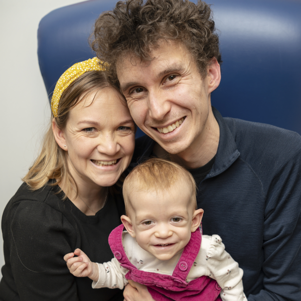 Parents Cameron and Dork sitting together in a large blue armchair with bay Safi, age 13 months, on their laps wearing pink dungarees. All three are smiling up into the camera.