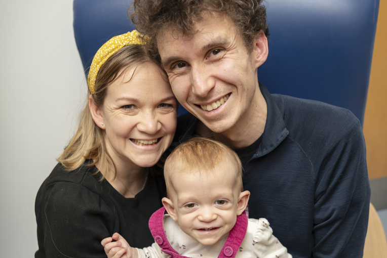 Parents Cameron and Dork sitting together in a large blue armchair with bay Safi, age 13 months, on their laps wearing pink dungarees. All three are smiling up into the camera.