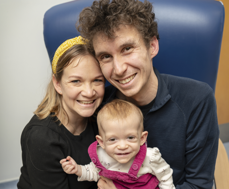 Parents Cameron and Dork sitting together in a large blue armchair with bay Safi, age 13 months, on their laps wearing pink dungarees. All three are smiling up into the camera.