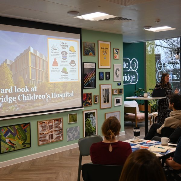 A woman standing in a room full of people presenting information about Cambridge Children's Hospital. There is a screen with an image of the new hospital on it
