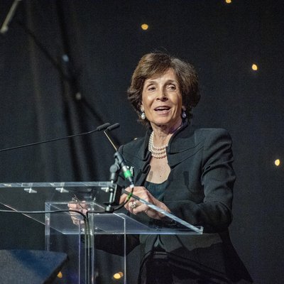 A woman with beautifully styled dark hair, wearing a dark suit and pearls, standing at a podium talking to an audience