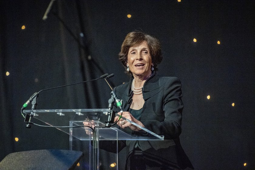 A woman with beautifully styled dark hair, wearing a dark suit and pearls, standing at a podium talking to an audience