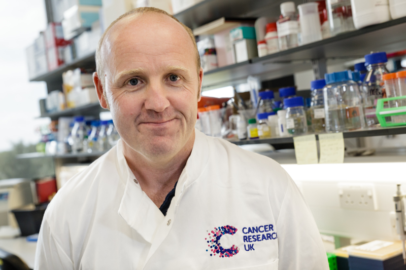 Dr Gill in a white lab coat in front of a lab bench and shelves full of bottles and boxes
