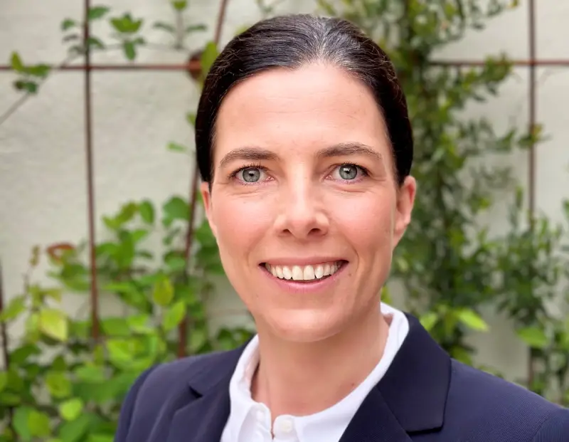 Dr Ellie Day wearing a dark suit jacket and white shirt smiling into the camera with a trellis with vines in the background.