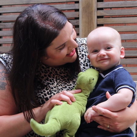 Mum Elaine sat on a bench entertaining baby Aidan with a toy tyrannosaurus