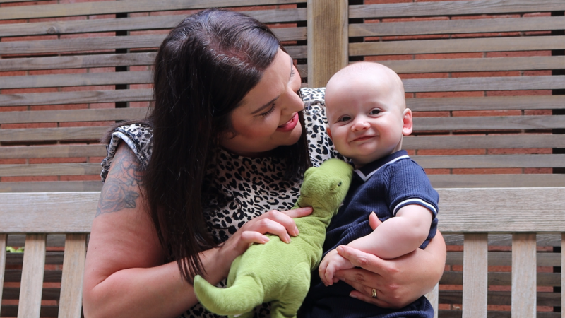 Mum Elaine sat on a bench entertaining baby Aidan with a toy tyrannosaurus
