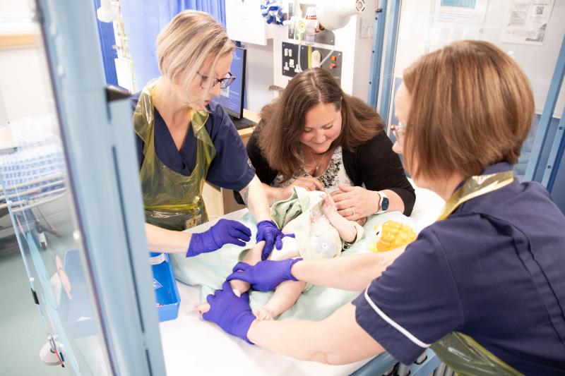Baby Elizabeth lying on a cot with her mum talking to her while a nurse holds her leg and another nurse prepares her for the COVID-19 vaccine as part of the diabetes prevention trial