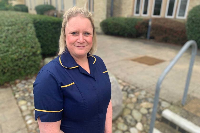A woman in navy nurses uniform standing outside. She has blond hair and is smiling