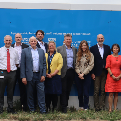 A group of people looking smart and smiling in front of the Cambridge Children's Hospital bright blue hoardings with the logo to the left