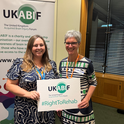 A woman with long brown hair and a patterned dress alongside a woman with short grey hair, glasses and a patterned dress. holding a sign saying #rightorehab
