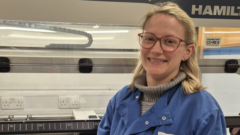 Emma standing in front of an automated liquid handling robot in the genetics lab wearing a blue lab coat