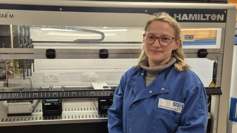 Emma standing in front of an automated liquid handling robot in the genetics lab wearing a blue lab coat
