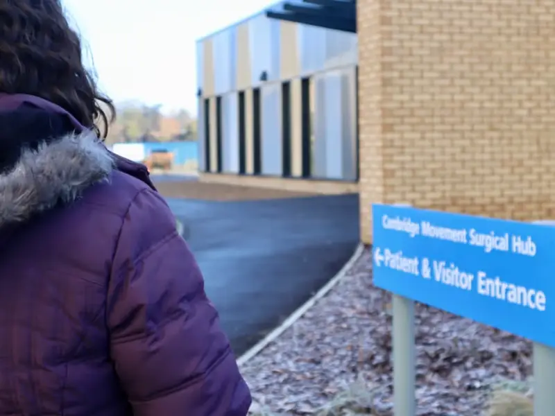 Person walking towards the patient and visitor entrance sign for Cambridge Movement Surgical Hub.