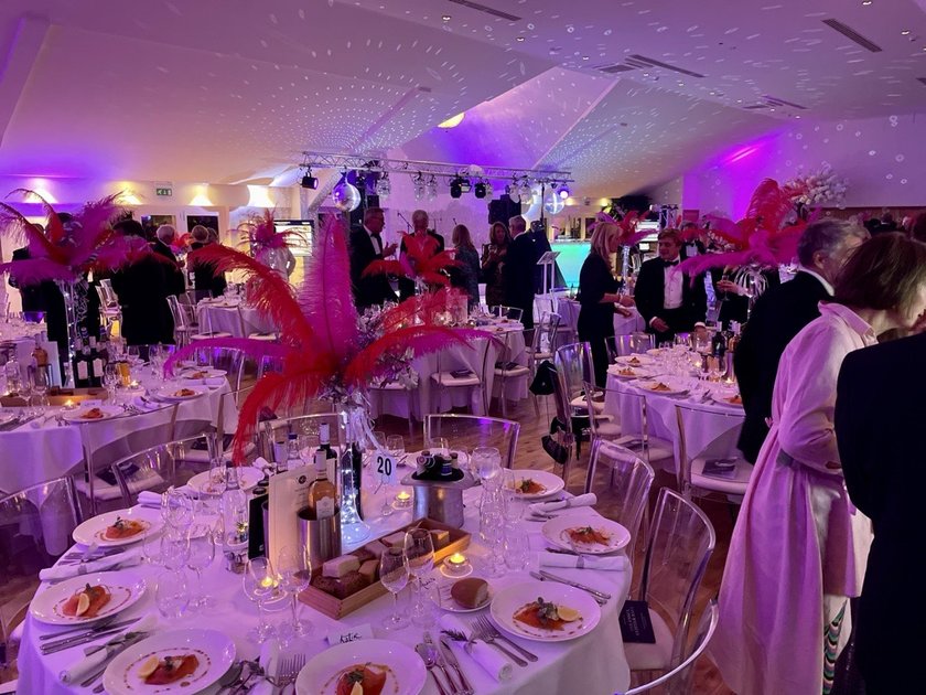 The inside of a marquee with round tables laid for a posh dinner with lots of people milling around in smart ball dresses and black tie suits