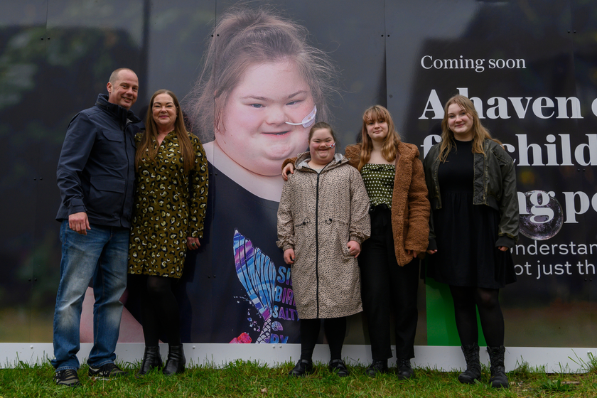 A family, with a mum and dad, plus three daughters, standing in front of a huge photo of one of the daughters. the photo is on the cambridge children's hospital hoardings