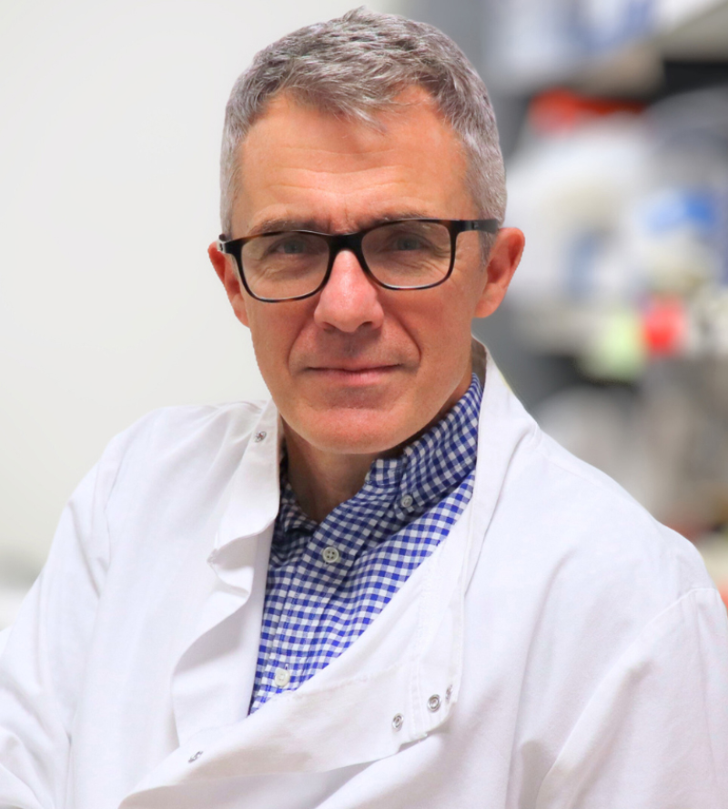 George Vassiliou wearing a blue check shirt under a white lab coat with lab shelves behind him
