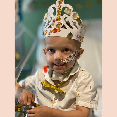 A little boy with big brown eyes wearing a paper crown with gold decorations and jewels on it. He is wearing a white shirt and a gold bow tie. He is smiling. He has a nasal tube and a tracheostomy tube.