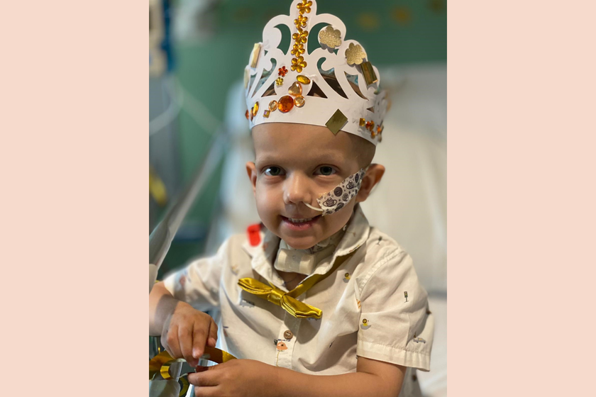 A little boy with big brown eyes wearing a paper crown with gold decorations and jewels on it. He is wearing a white shirt and a gold bow tie. He is smiling. He has a nasal tube and a tracheostomy tube.
