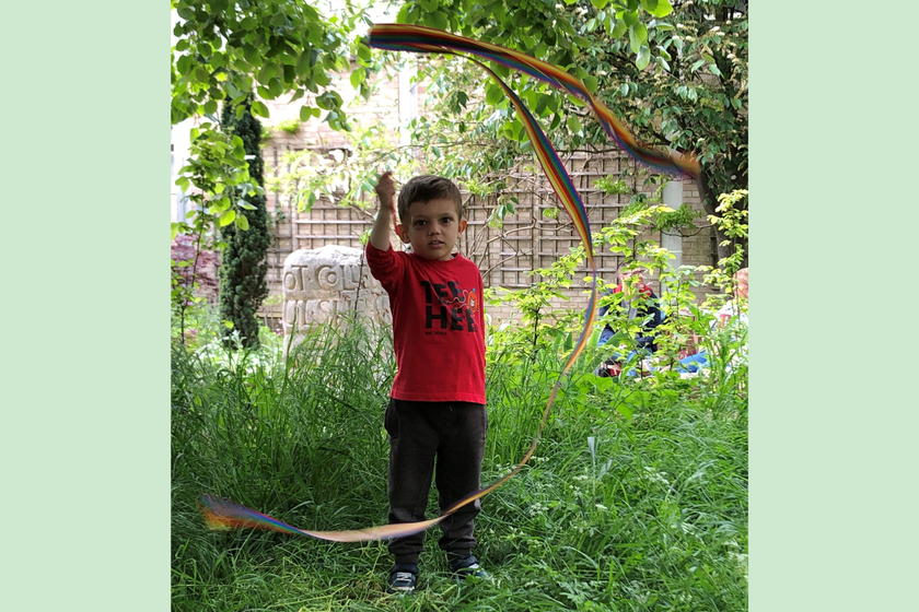 A little boy with brown hair, in a red t shirt, standing in long grass under a tree. He is waving a swirly ribbon above his head