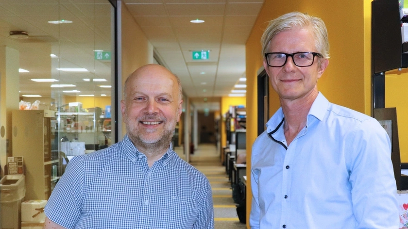 Bertie and Matthias standing together outside their labs in Cambridge. They are both wearing blue button down shirts and smiling into the camera.