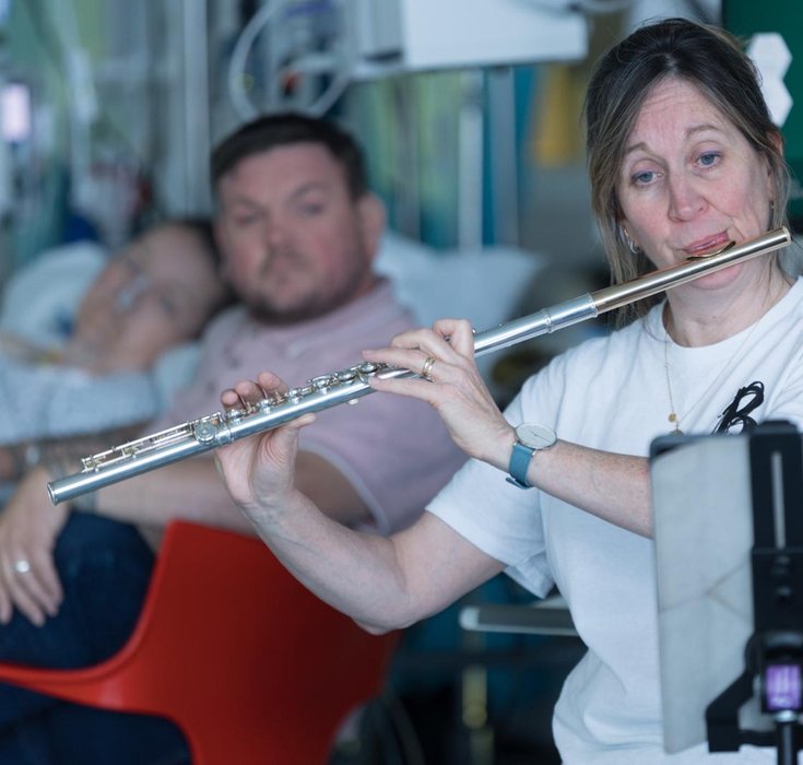 A Patient and visitor listen to a Britten Sinfonia Musician play the flute on a hospital ward.