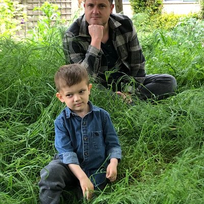 A young boy sitting in long grass with his dad sitting behind him