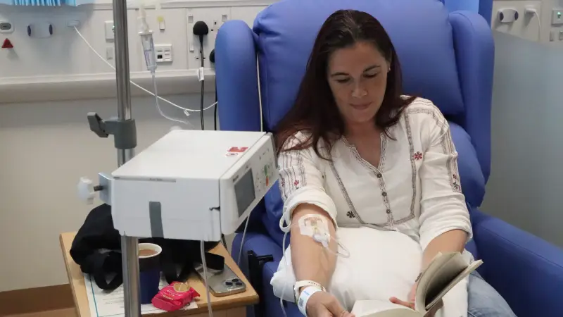 Isobel sat in a blue padded arm chair reading a book with a drip in her right elbow. The drip stand with the infusion bag is in the foreground.