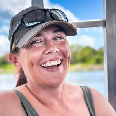 Isobel smiling into the camera wearing a green vest top, and peaked cap with sunglasses on the brim. The background shows water, a green coastline and blue clouded sky.
