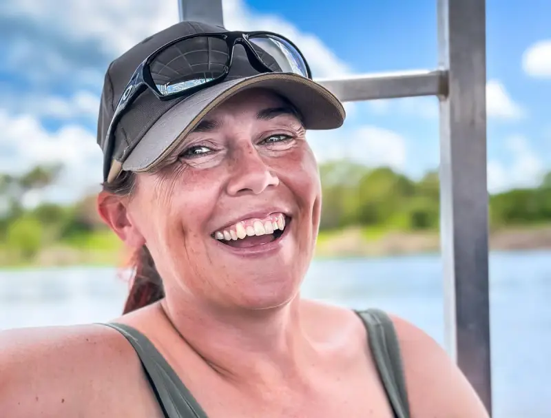 Isobel smiling into the camera wearing a green vest top, and peaked cap with sunglasses on the brim. The background shows water, a green coastline and blue clouded sky.