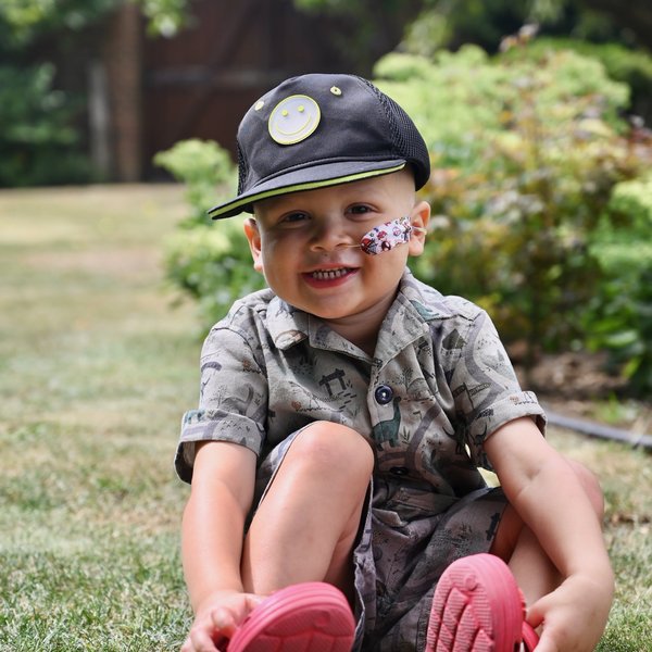 Jacob, a young boy wearing red crocs a dinosaur print outfit and black cap. He has a nasogastric tube taped to his left cheek and is sat on a lawn in the garden smiling into the camera.