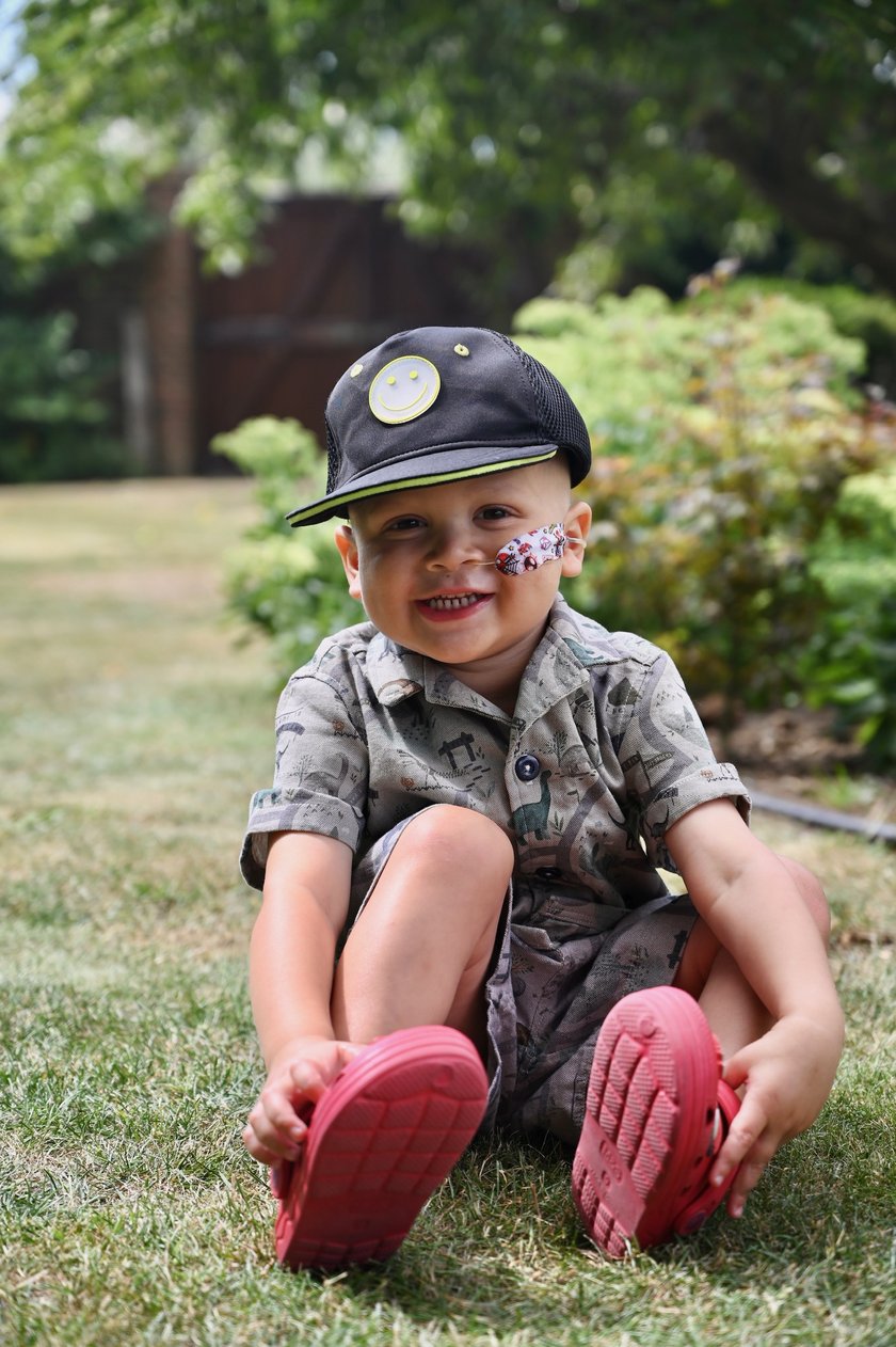 Jacob, a young boy wearing red crocs a dinosaur print outfit and black cap. He has a nasogastric tube taped to his left cheek and is sat on a lawn in the garden smiling into the camera.